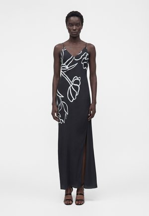 Femme portant une robe maxi noire sans manches avec contour floral blanc, col en V, fente latérale et sandales noires à lanières, debout sur fond blanc.