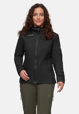 Frau mit lockigem Haar trägt eine schwarze Jacke mit Reißverschluss und olivgrüne Hosen und steht vor einem schlichten hellen Hintergrund.