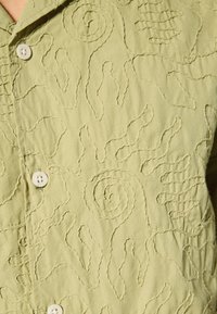 Lichtgroen overhemd met knopen en textuur van borduurwerk met krullen en organische vormen, gemaakt van zacht stof.