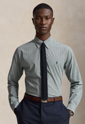 Homme portant une chemise grise à carreaux boutonnée, cravate tricotée foncée, ceinture marron, pantalon bleu marine, mains dans les poches sur fond uni.