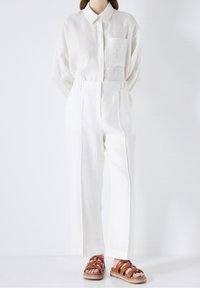 Vit linneskjorta med ficka, parat med högmidjade vita byxor. Bruna lädersandaler med flera remmar.