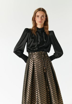 Schwarze Satin-Bluse mit langen Ärmeln und gerafften Schultern, kombiniert mit einem Rock im goldenen Zickzackmuster, der einen Kordelzugbund und Quasten hat.