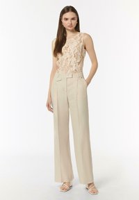 Ärmelloses beiges Oberteil mit floralem Spitzenmuster, kombiniert mit hoch taillierten, weit geschnittenen cremefarbenen Hosen. Weiße Riemchenschuhe runden das Outfit ab.