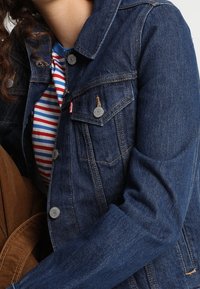 Giacca in denim blu scuro con taglio classico, caratterizzata da bottoni argentati, due tasche sul petto e cuciture visibili. Camicia a righe sotto.