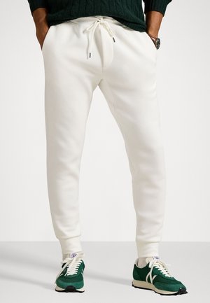 Pantalones de chándal blancos con cintura ajustable, que presentan una textura suave y piernas ajustadas. Llevados con zapatillas verdes y un suéter verde oscuro.