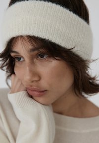 Gruba, biała dziana opaska na głowę o miękkiej fakturze, noszona z beżowym swetrem. Zbliżenie twarzy pokazujące naturalny odcień skóry i brązowe oczy.