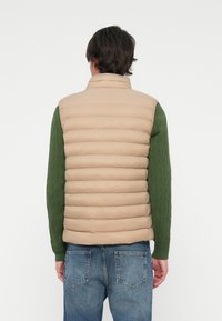 Beige padded vest with a high collar, features horizontal stitching and a smooth texture, worn over a green cable-knit sweater. Denim jeans visible.