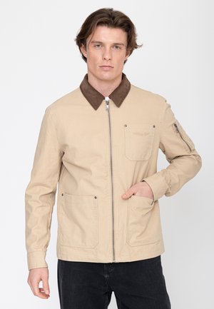 Man draagt een beige jas met ritssluiting, een bruine kraag, voorzakken en een klein ritszakje op de linkermouw, poserend tegen een witte achtergrond.