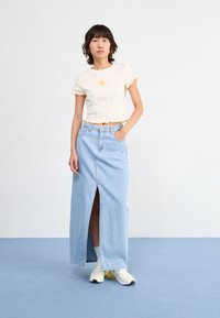 Vit ribbad crop top med gula vågiga kanter och små blommiga broderier, i kombination med en ljusblå lång jeanskjol med en framspalt.