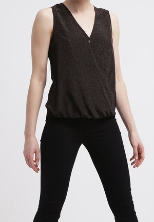 Vrouw draagt een donker patroonloze mouwloze V-hals top met knoop en nauwsluitende zwarte broek, staat met ontspannen armen.