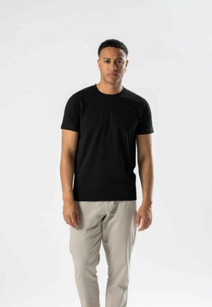 Mand med kort hår iført en sort t-shirt med korte ærmer og lyse beige bukser, stående mod en ensfarvet hvid baggrund.