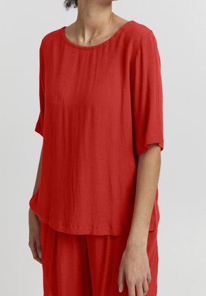 Kvinde iført løs, rød tekstureret kortærmet top og matchende bukser, stående mod en ensartet lysegrå baggrund.