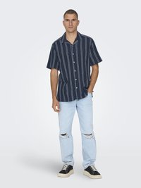 Chemise bleu marine à manches courtes avec des rayures blanches, à boutonnière. Portée avec un jean bleu clair déchiré et des baskets noires avec des semelles blanches.