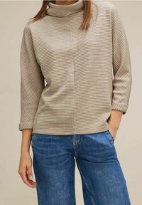 Beige randig tröja med hög krage och trekvartsärmar. Tyget verkar mjukt och matchas med blå vida jeans.