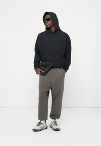 Schwarzer Kapuzenpullover, anthrazitfarbene tapered Pants und beige Sportschuhe. Accessoires umfassen eine kleine schwarze Gürteltasche und dunkle Sonnenbrillen.