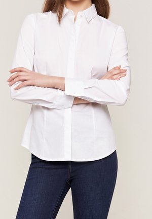 Camicia bianca a maniche lunghe con colletto, caratterizzata da una texture liscia e un design aderente, abbinata a jeans blu scuro.