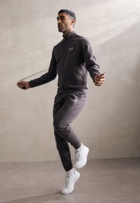 Completo sportivo grigio con giacca con zip e pantaloni; modello che salta la corda; sneakers bianche; design minimalista con branding sottile.