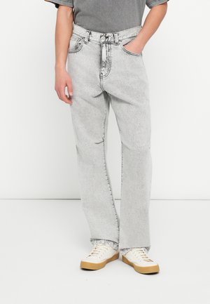 Inimene, kes kannab helehalli happepesuga teksapükse, valgeid tossusid kummist tallaga ja halli lühikese varrukaga särki, seisab valge tausta ees.