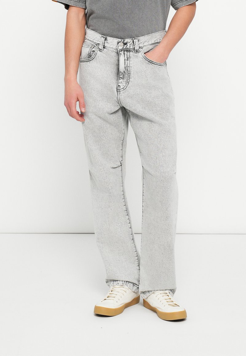 Άτομο που φορά ανοιχτό γκρι τζιν με όξινη πλύση, λευκά αθλητικά παπούτσια με καουτσούκ σόλες και γκρι κοντομάνικη μπλούζα, στέκεται μπροστά σε λευκό φόντο.