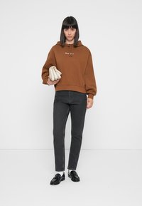 Brun hettegenser med en avslappet passform, sammen med grå slim-fit jeans. Svarte loafers og en kremfarget håndveske fullfører looken.