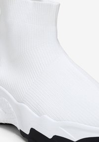 Zapatilla alta de punto blanca con textura acanalada. Presenta una suela negra gruesa con un diseño ondulado y una parte superior tipo calcetín sin costuras.