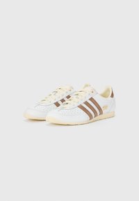 Adidași albi cu dungi de piele maro și accente de fildes. Caracteristici: design perforat, vârf rotund și talpă din cauciuc. Detaliu cu logo aurit pe lateral.