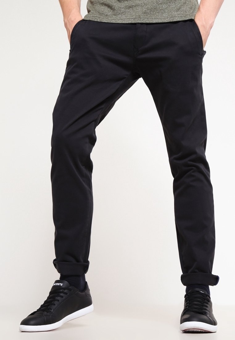 Černé kalhoty slim fit, vyrobené z hladkého materiálu. Mají boční kapsy a ohrnuté rukávy, doplněné o černé tenisky.