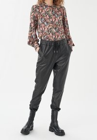 Blouse à motifs colorés avec des manches longues et un col à ouverture en forme de clé, accompagnée de pantalons jogger en cuir noirs et de bottines noires.