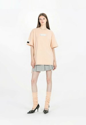 Joven mujer con camiseta de color melocotón de gran tamaño, falda plisada gris, calcetines anchos melocotón y tacones negros, posando sobre fondo blanco.