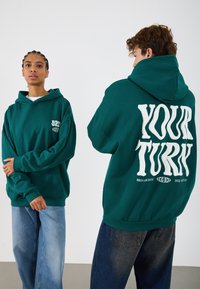Türkisfarbener, übergroßer Hoodie mit Logo auf der Vorderseite, großem weißen Text auf dem Rücken, gerippten Bündchen und Saum, kombiniert mit lockeren, hellen und dunklen Jeans.