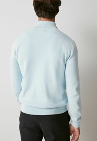 Šviesiai mėlynas megztinis su aukštu apykakle, raukšlėtais rankogaliais ir apačia, pagamintas iš minkštos, tekstūruotos megztos medžiagos.