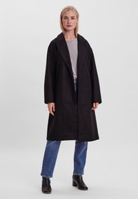 Cappotto nero lungo con colletto a risvolto, vestibilità ampia e tasche laterali, indossato sopra un top grigio e jeans blu, abbinato a stivali neri alla caviglia.