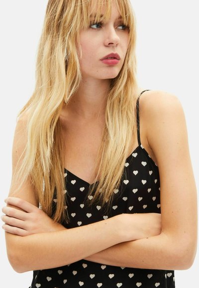 Schwarzes Camisole mit weißem Herzmuster, dünnen Trägern, V-Ausschnitt und leichtem Stoff, mit entspannter Passform und subtiler Textur.