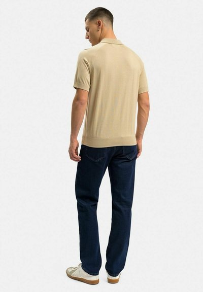 Beige kortärmad polo med en subtil textur, kombinerad med mörkblå raka jeans och ljusa sneakers.