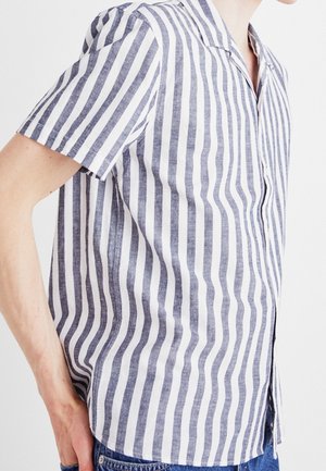 Persoon die een overhemd met korte mouwen met verticale witte en blauwe strepen en een blauwe spijkerbroek draagt, zichtbaar vanaf het midden van de romp tot de nek.