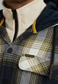 Chaqueta a cuadros en blanco, oliva y marino con collar con cremallera. Presenta botones negros, un acento amarillo en la capucha y una etiqueta PME LEGEND.