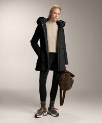 Czarna puchowa kurtka z kapturem wykończonym futrem, beżowy prążkowany golf, czarne legginsy, brązowe trampki i brązowa torba na ramię.