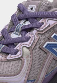 Gros plan d'une basket New Balance avec des lacets violets sur un tissu texturé beige et rose et un détail du logo bleu sur le côté.