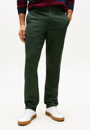 Man in een donker groene broek, witte sneakers met bruine zolen, en een donkere trui met rode en marineblauwe strepen, met zijn handen in zijn zakken.
