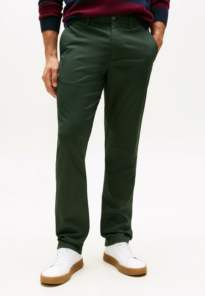 Man in een donker groene broek, witte sneakers met bruine zolen, en een donkere trui met rode en marineblauwe strepen, met zijn handen in zijn zakken.