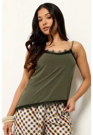 Kvinde med langt mørkt hår iført en olivengrøn camisole med sort blondekant og ternede bukser, poserer med den ene hånd på hoften og hvide armbånd.