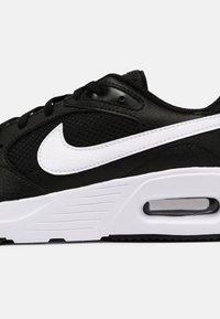Black sneaker with mesh and textured leather upper, white Nike swoosh, padded collar, and visible air cushioning in the white sole.