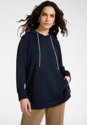 Donna con capelli lunghi e scuri, indossa una felpa con cappuccio blu navy con tasca frontale e pantaloni beige, in piedi contro uno sfondo chiaro.