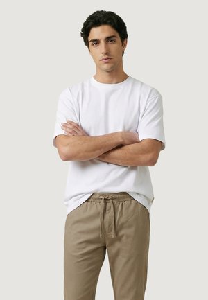 Giovane uomo con capelli scuri, che indossa una camicia bianca a maniche corte e pantaloni beige con coulisse, in piedi con le braccia incrociate.