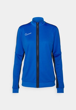 Blaues Sportjacke mit hohem Kragen, durchgehendem Reißverschluss, schwarzen Seiteneinsätzen und weißem Nike-Logo auf der linken Brust. Glatte Stoffstruktur.
