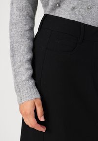 Svarta högmidjade byxor med slät textur, försedda med en sidoficka och midjeband, i kombination med en ljusgrå stickad tröja.