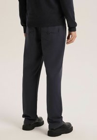 Granatowe spodnie o prostym kroju, wyposażone w płaskie tylne kieszenie i teksturowaną tkaninę. Noszone z czarnymi butami na masywnej podeszwie.