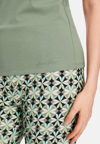 Olivgrünes Hemd mit glatter Textur, kombiniert mit gemusterten schwarzen Leggings, die grüne und weiße Blumenmuster aufweisen. Dezentes Markenlogo am Saum des Hemdes.