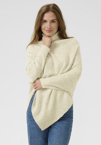 Tunic tricotat de culoare crem, cu guler înalt, având un model texturat în zigzag și o margine asimetrică. Purta cu blugi albaștri.