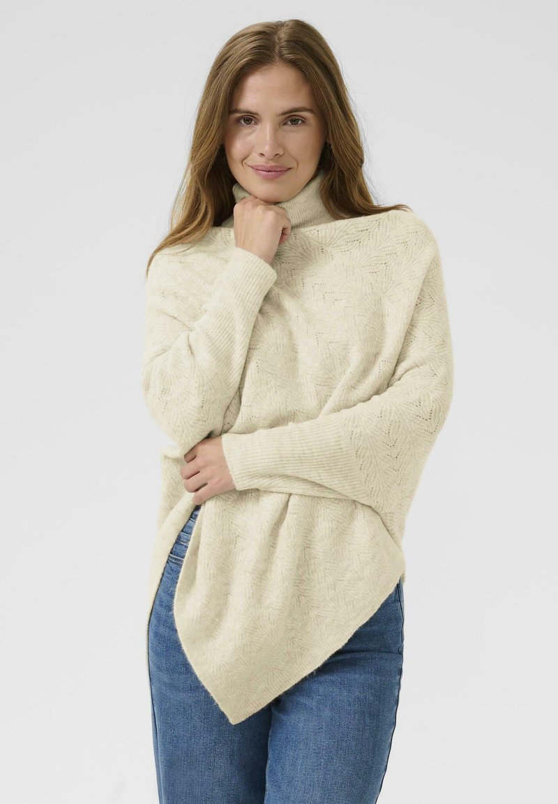 Tunic tricotat de culoare crem, cu guler înalt, având un model texturat în zigzag și o margine asimetrică. Purta cu blugi albaștri.